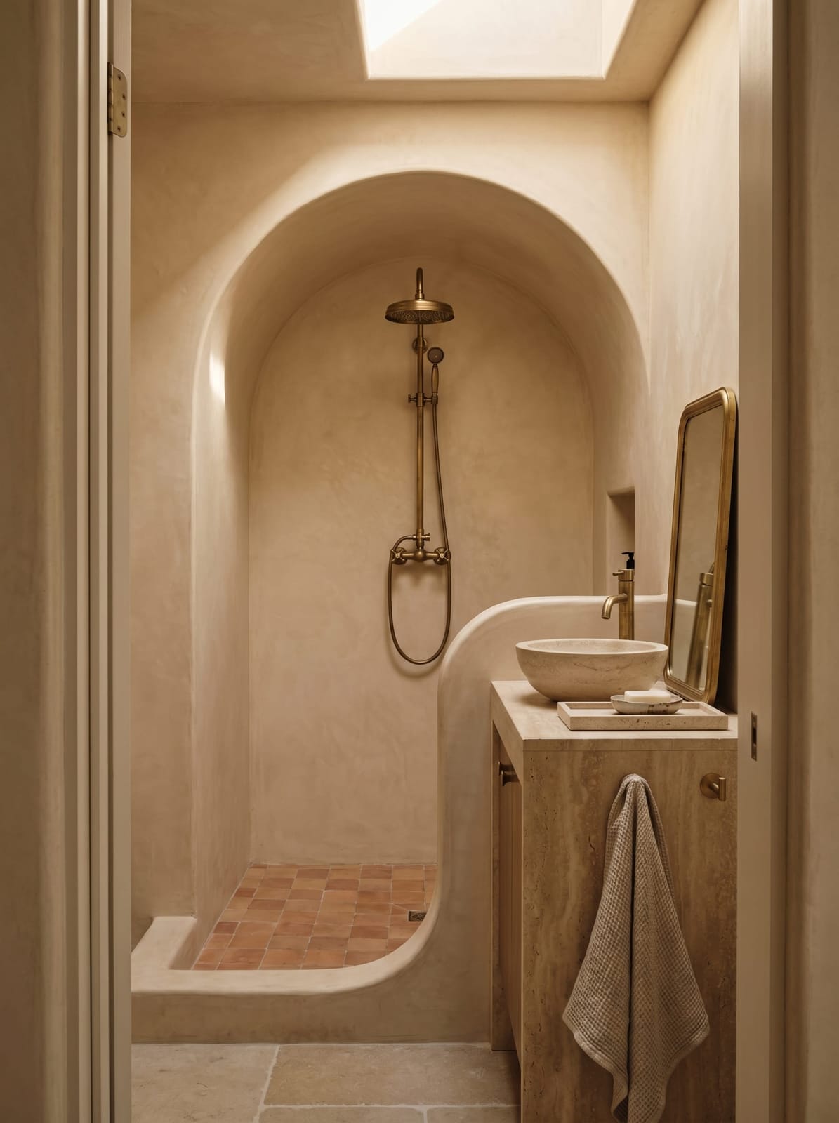 Warm organic bathroom with a curved lime plaster arch walk-in shower, handmade unglazed terracotta floor tiles, travertine sintered stone vanity with white oak drawers, round travertine vessel sink, brushed gold wall-mounted faucet with knurled handles, rounded rectangle brass mirror, and antique beige waffle-weave bath towels.