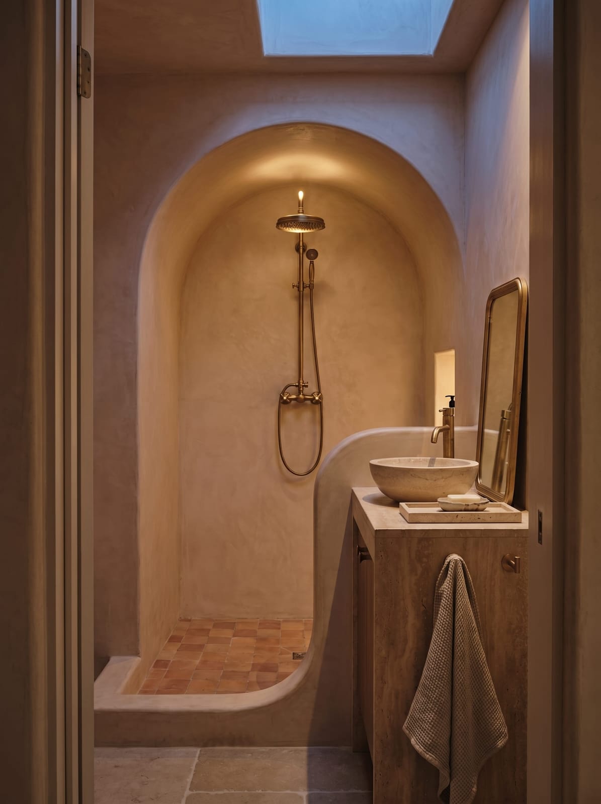 Arched lime plaster walk-in shower and travertine vanity with brushed gold fixtures, shown under natural skylight and warm dusk ambiance.