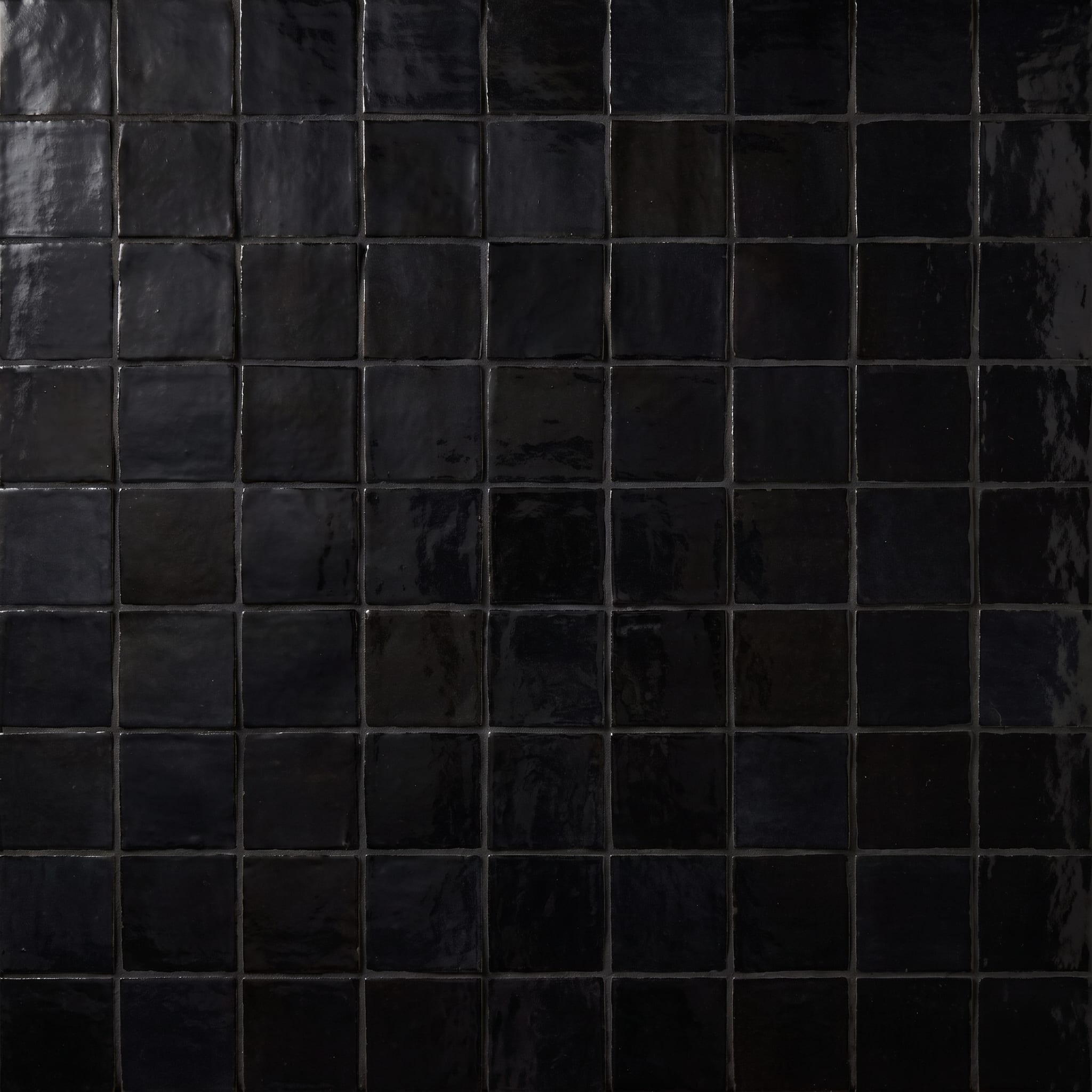 Close-up of glossy black square Zellige-style tile showing uneven glazed surfaces catching warm directional light.