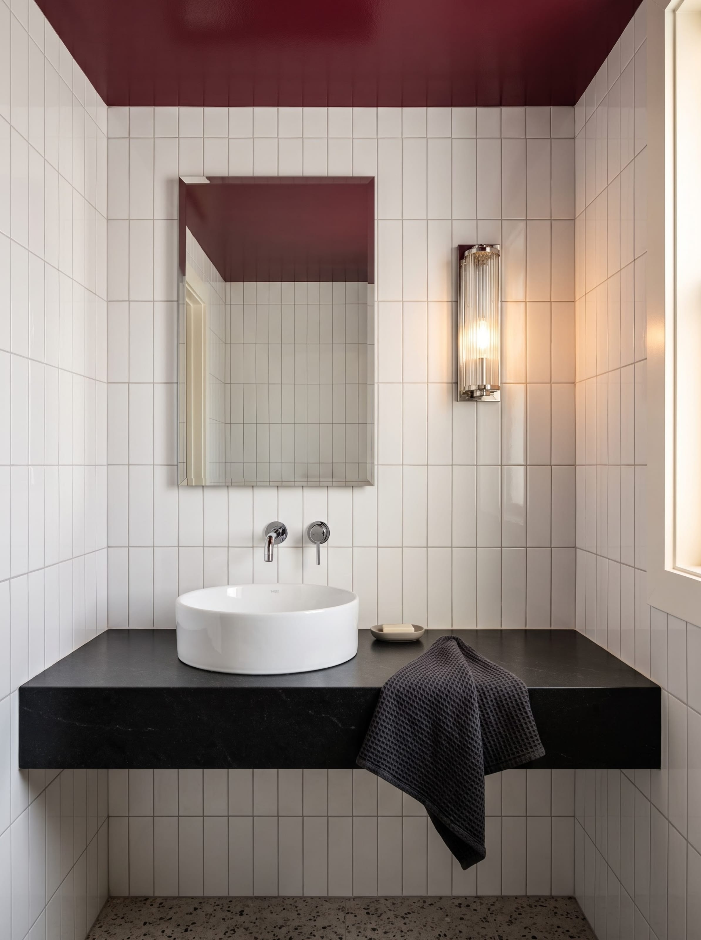 Compact powder room design study with a glossy burgundy ceiling and white vertical tile shown under daylight and evening sconce lighting.