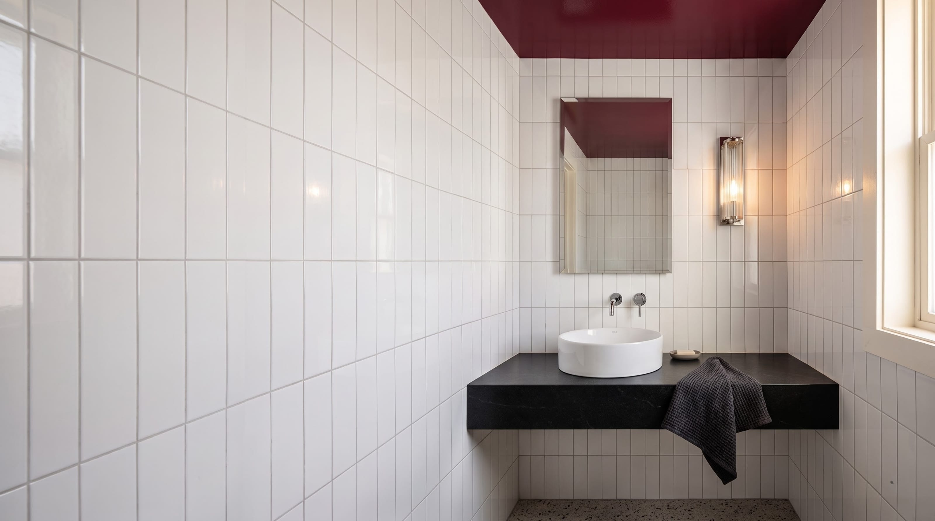 A compact powder room featuring a high-gloss burgundy ceiling, vertically stacked white subway tile, a thin black floating counter, and a round white vessel sink in soft natural daylight.