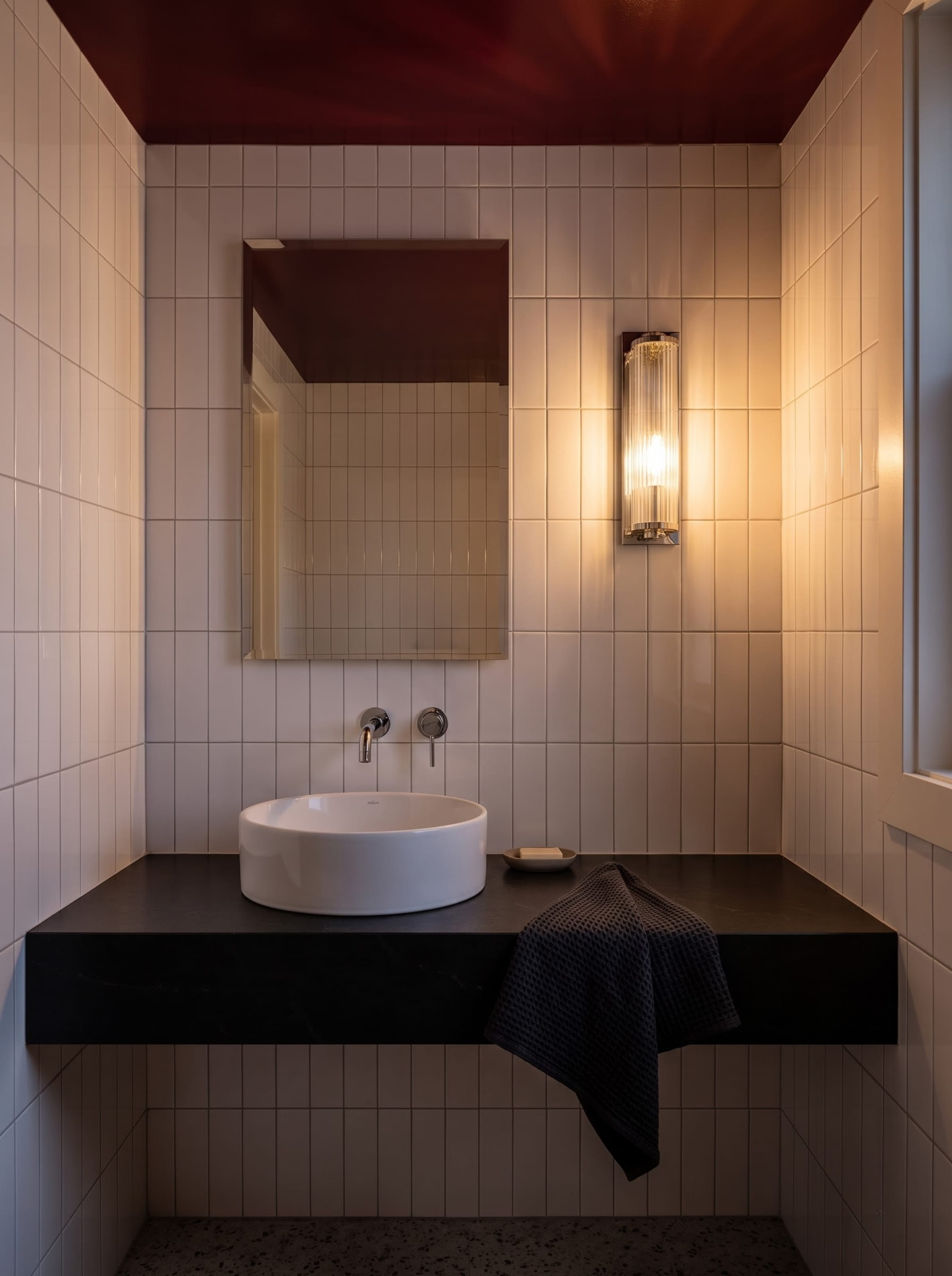 Compact powder room design study with a glossy burgundy ceiling and white vertical tile shown under daylight and evening sconce lighting.