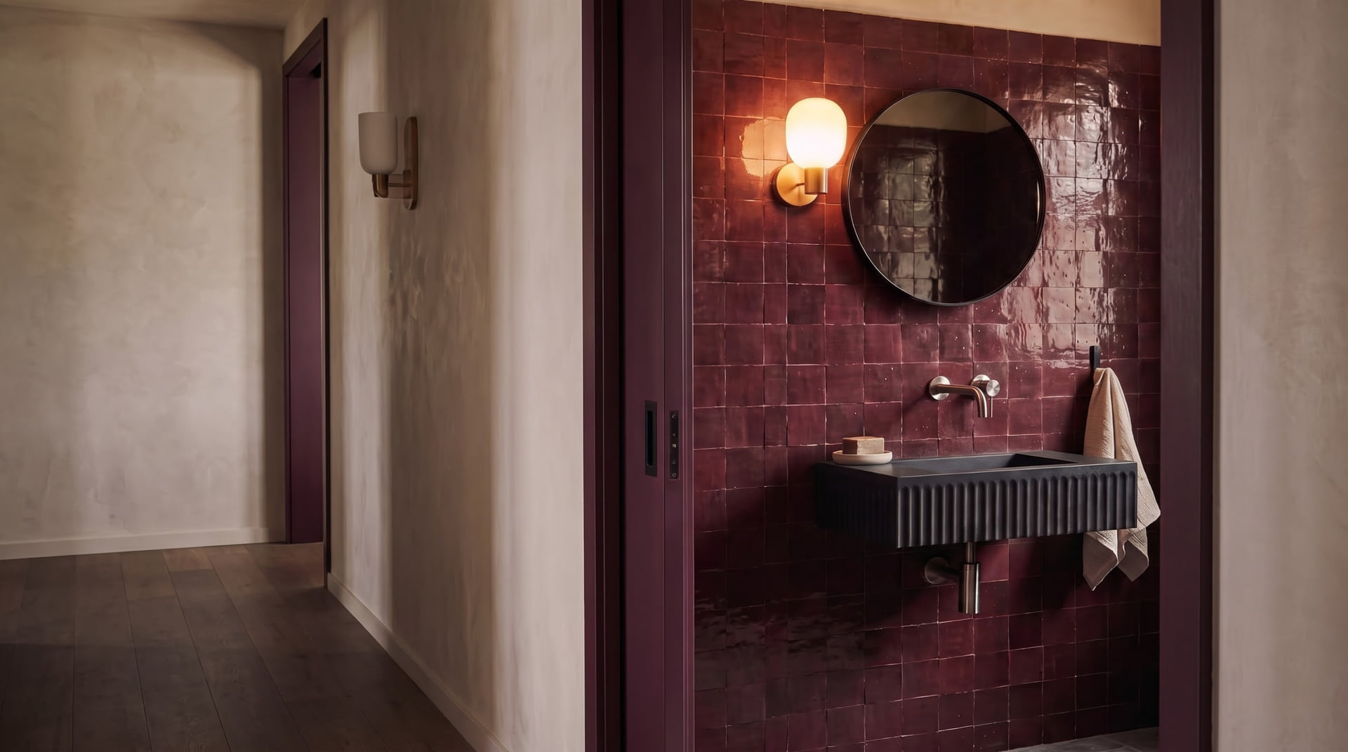 Wide perspective of a burgundy powder room design study with glossy maroon tile, brass sconces, black round mirror, and black wall basin under night sconce lighting.