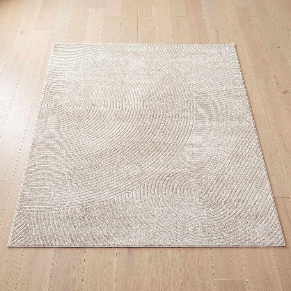Beige low-pile area rug with a carved concentric ripple pattern on a light wood floor
