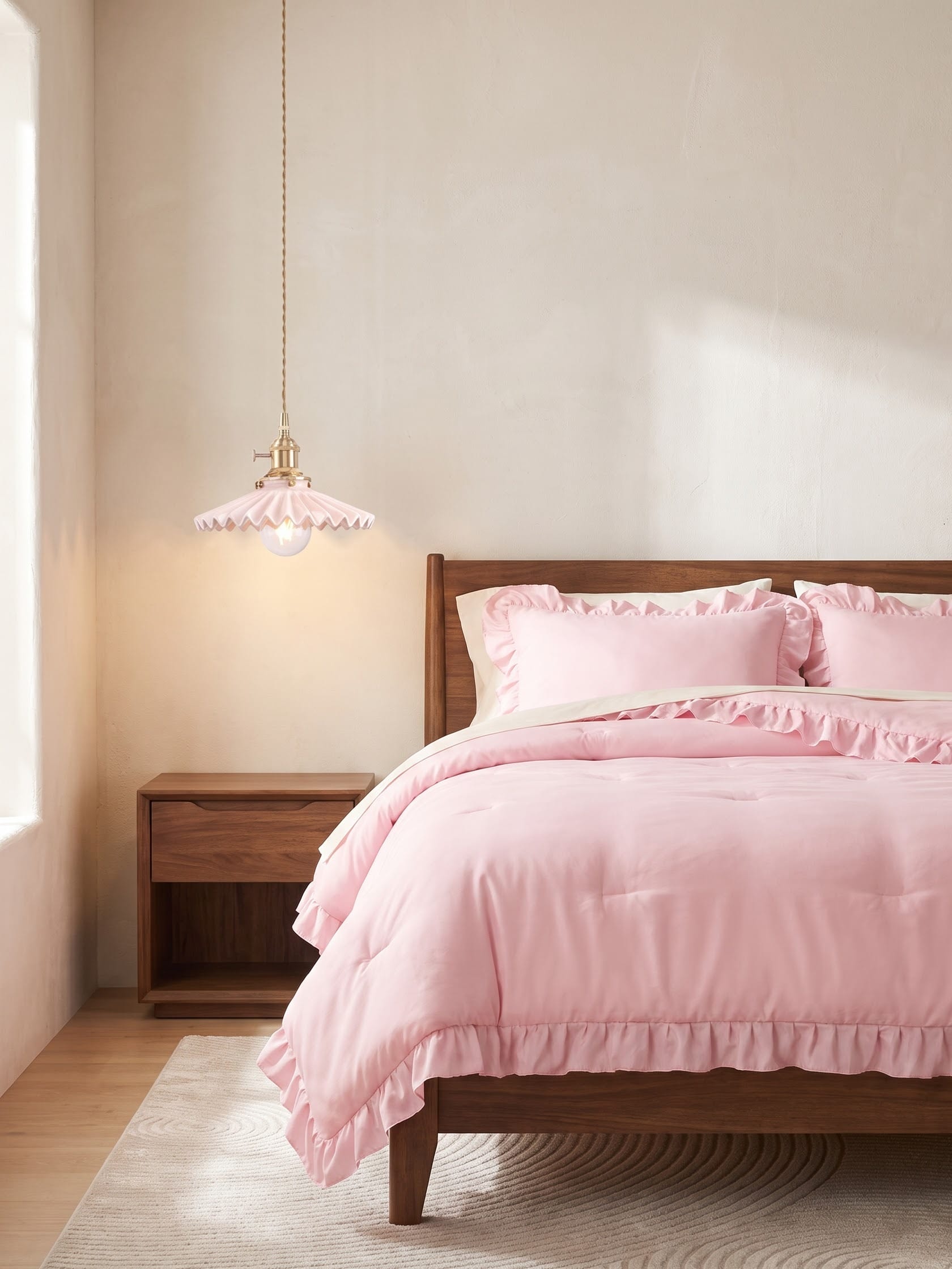 Blush pink ruffled bedding on a walnut bed beside a walnut nightstand, with a pink pleated pendant on a brass fitting and a beige rug.