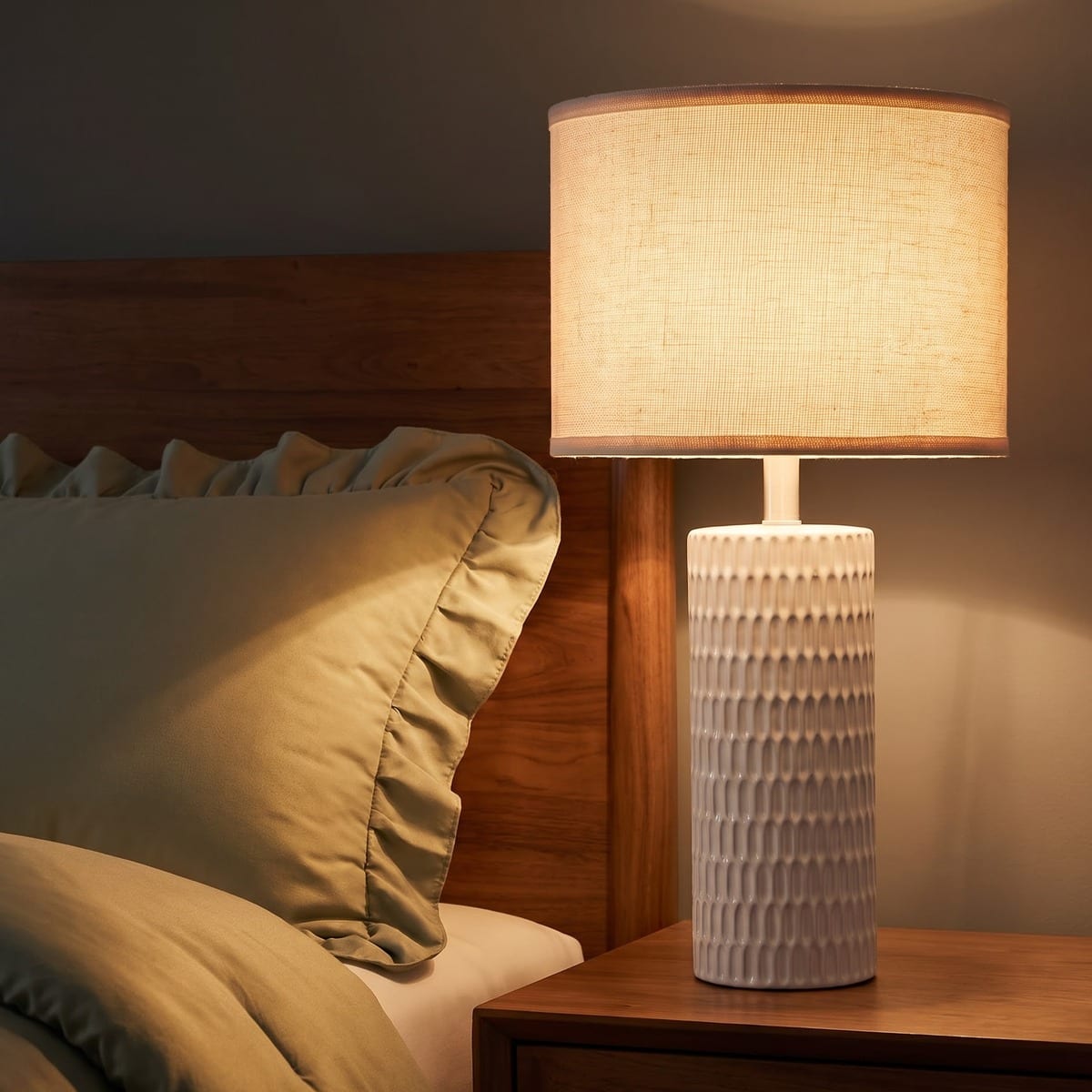 Lit white carved ceramic table lamp with a linen drum shade beside a sage ruffled pillow on a walnut nightstand at night.