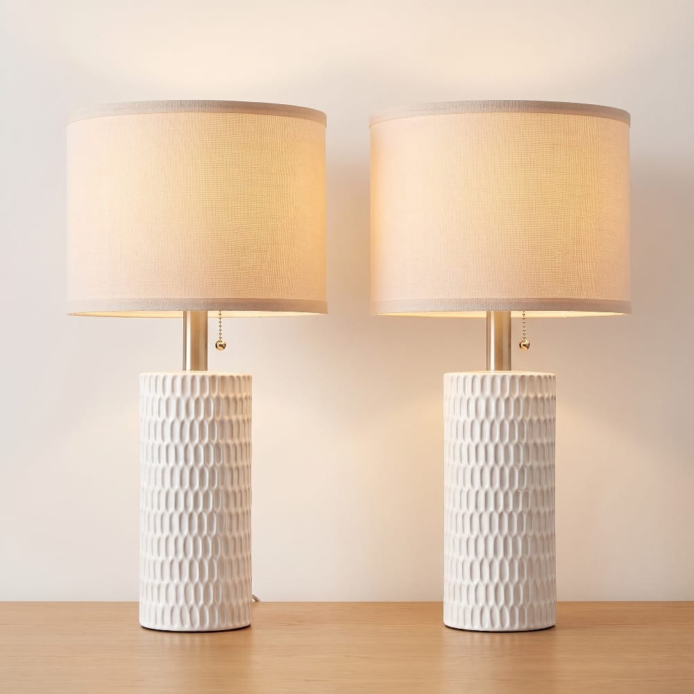 Pair of white textured ceramic table lamps with linen drum shades lit on a wood surface