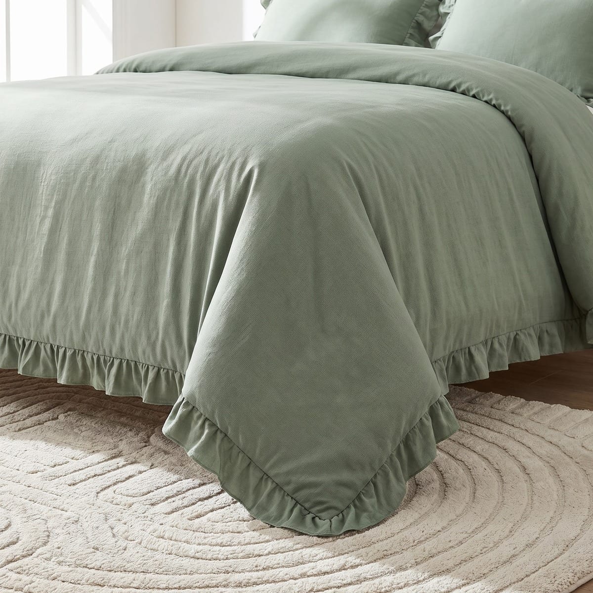 Close-up of a sage green ruffled comforter corner falling onto a beige rug with a carved concentric pattern.