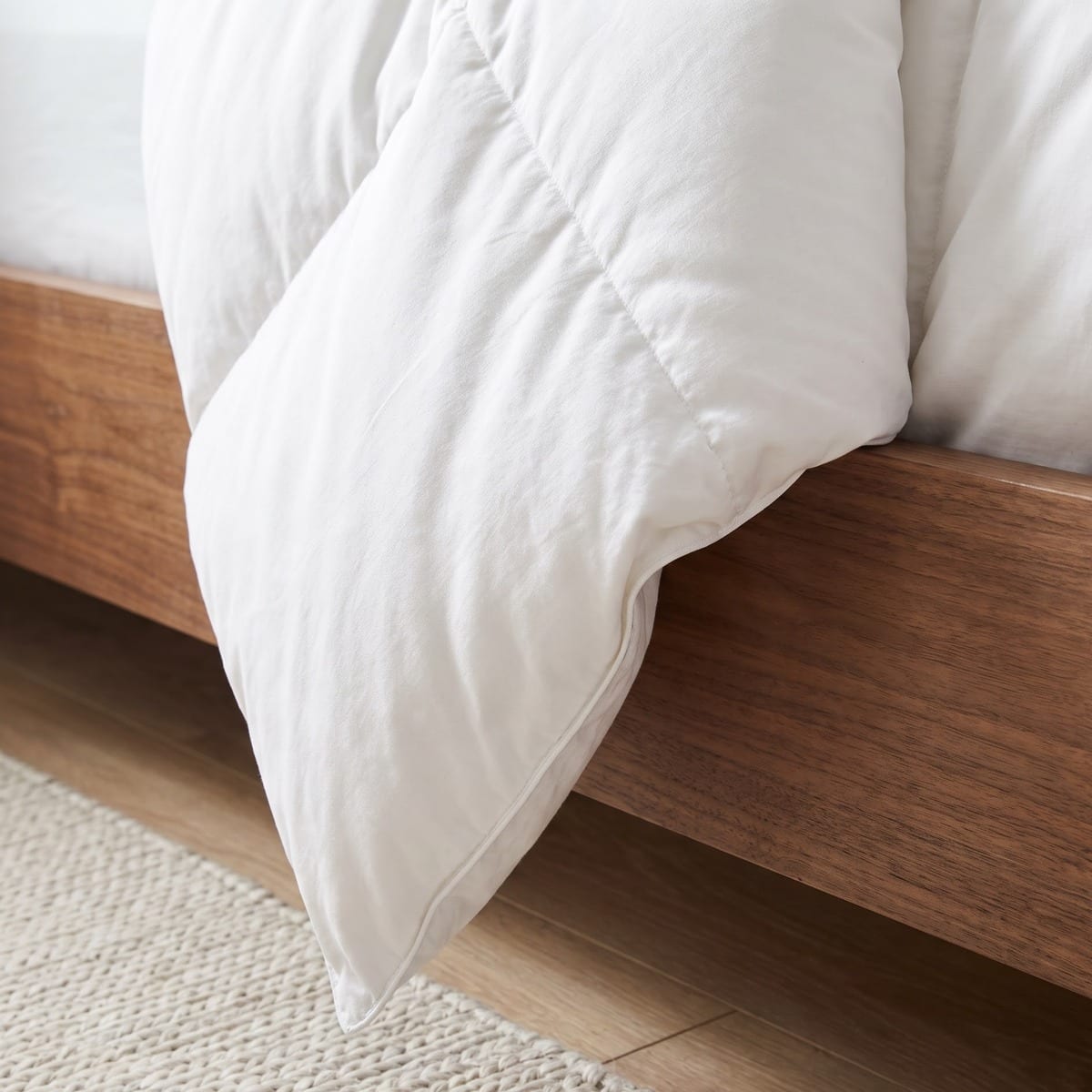 Corner of a white quilted comforter draped over a walnut bed frame above a woven cream rug.
