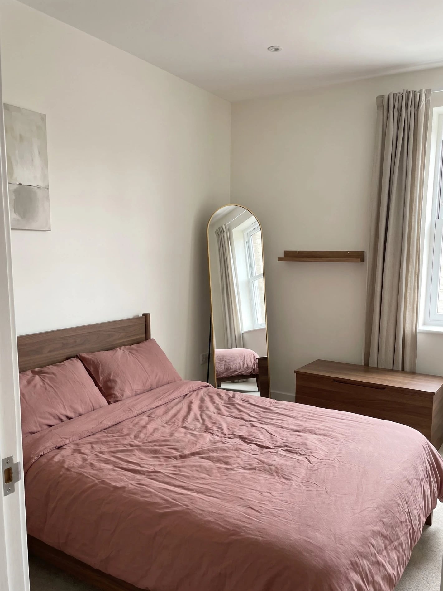 Compact bedroom featuring wood storage, soft rose bed linen, and an arched full-length mirror