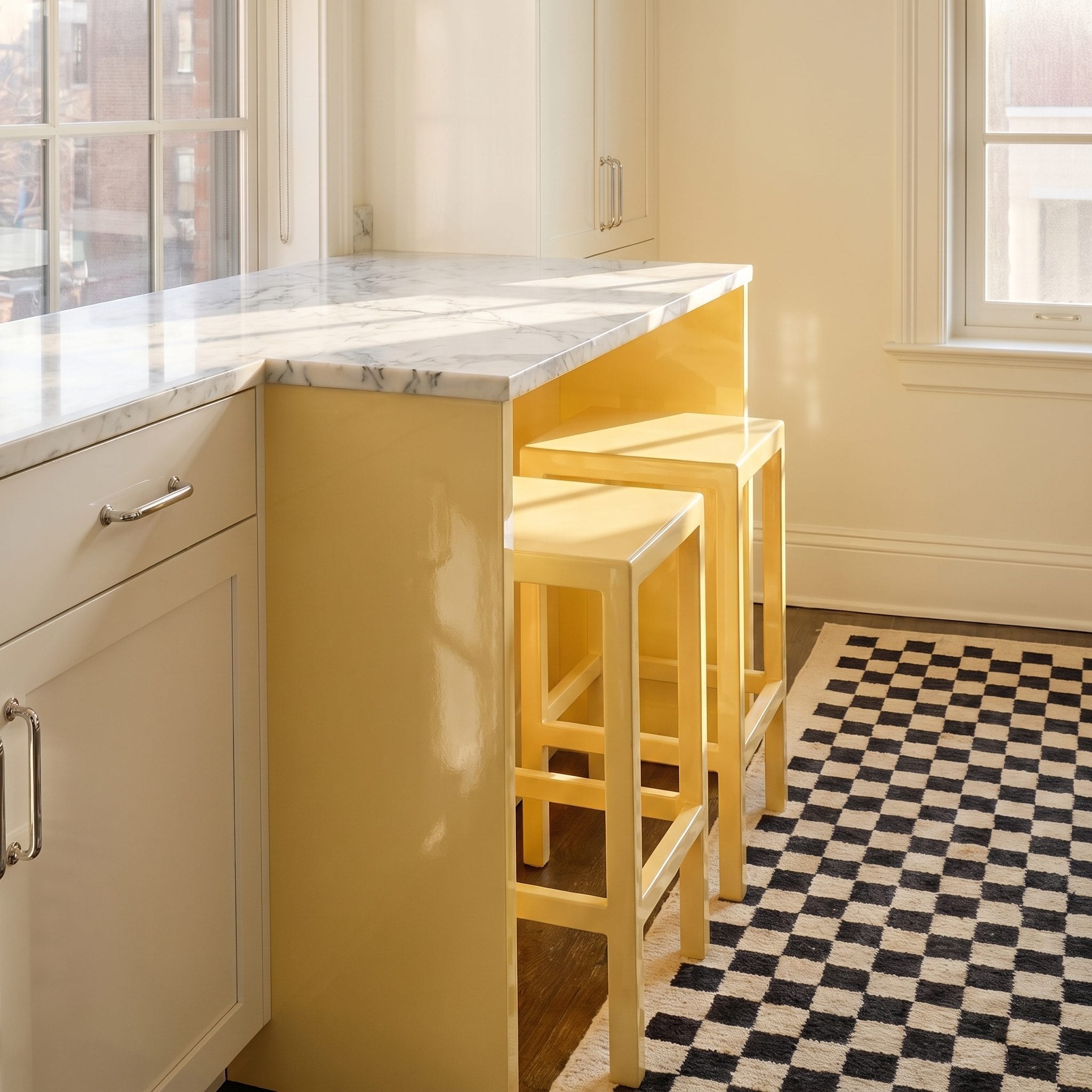 Pale yellow backless counter stools beside a butter yellow marble kitchen peninsula and checkerboard floor.
