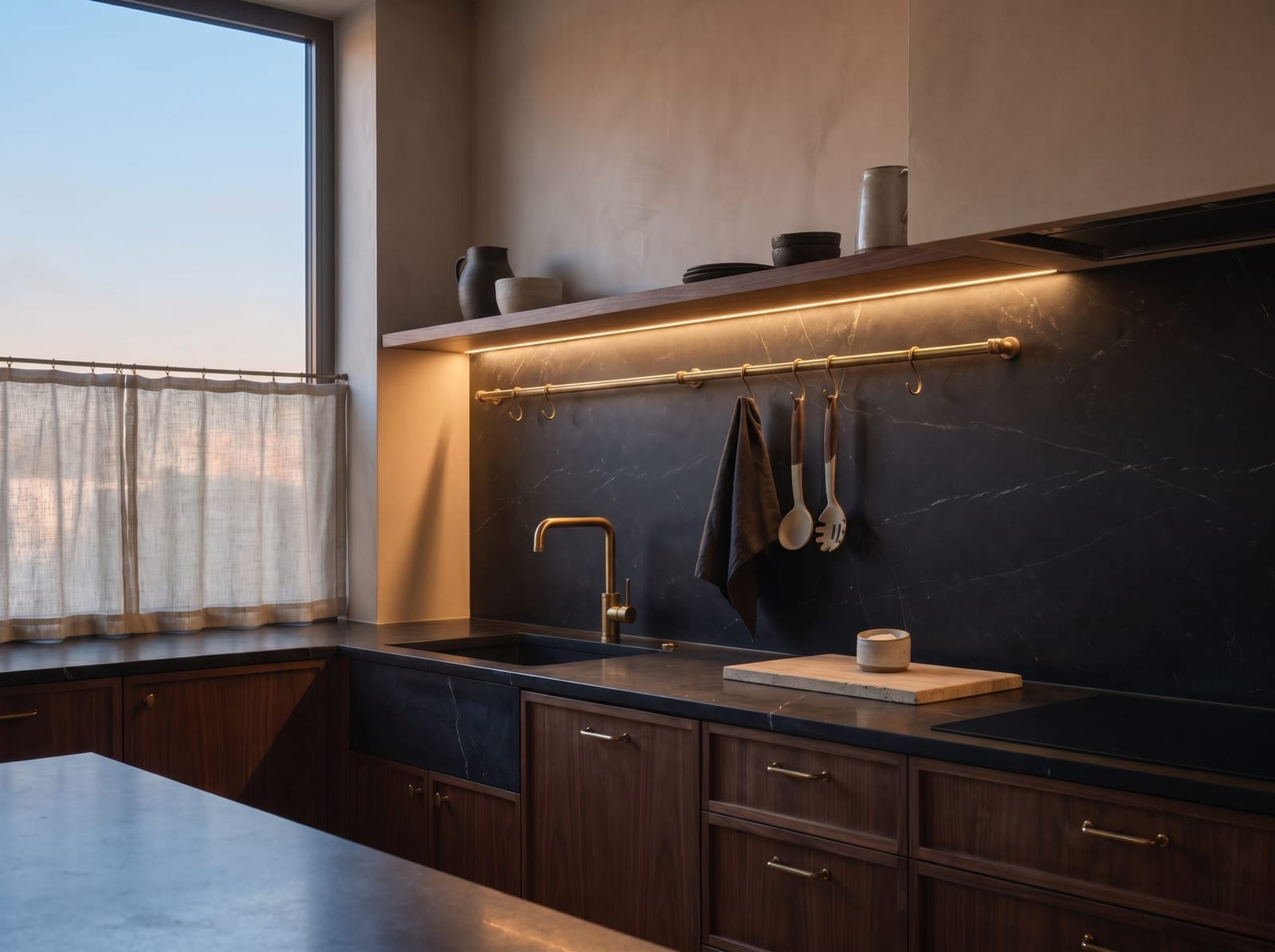 A horizontal close-up view of the moody kitchen showing the brushed brass faucet, matte black farmhouse sink, unlacquered brass pot rail, and edge-grain teak cutting board on the dark soapstone counter.