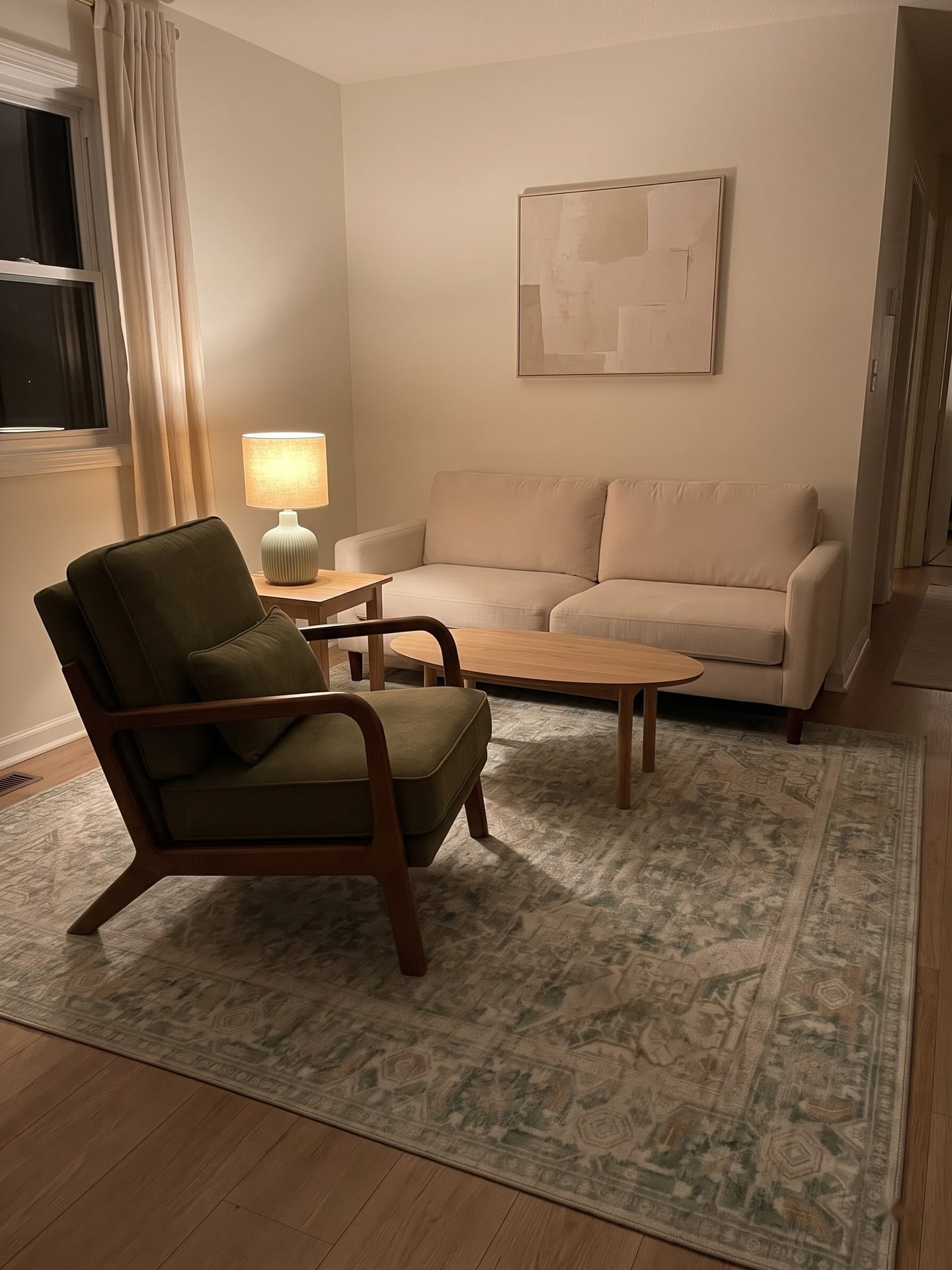 Warm living room in evening light with a green armchair opposite a beige sofa on a pale patterned rug with a lit celadon lamp.