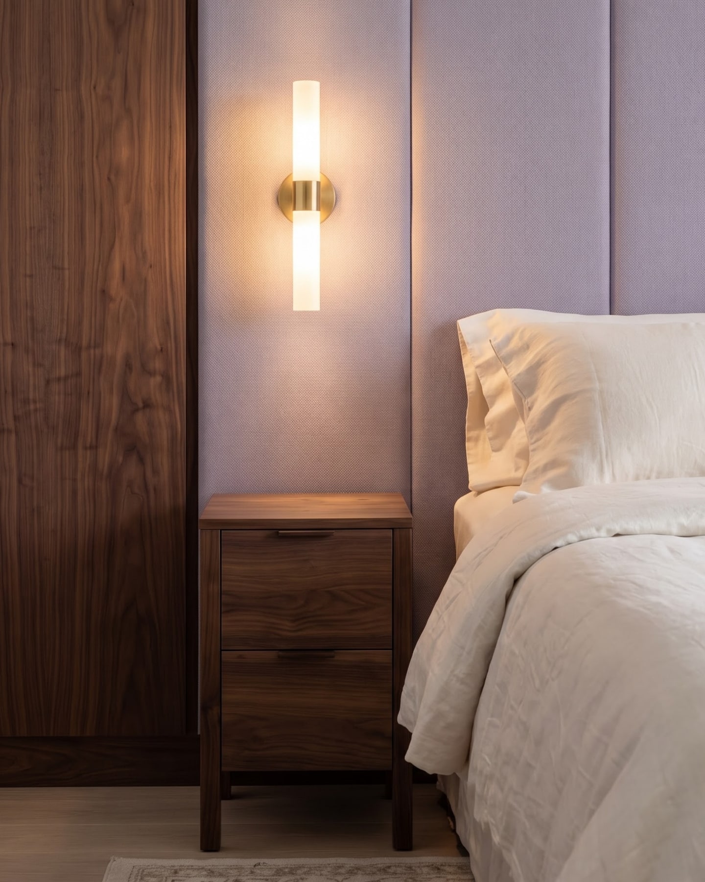 Bedside arrangement featuring a walnut two-drawer nightstand, dual-ended glowing sconce, and lilac fabric wall panel.