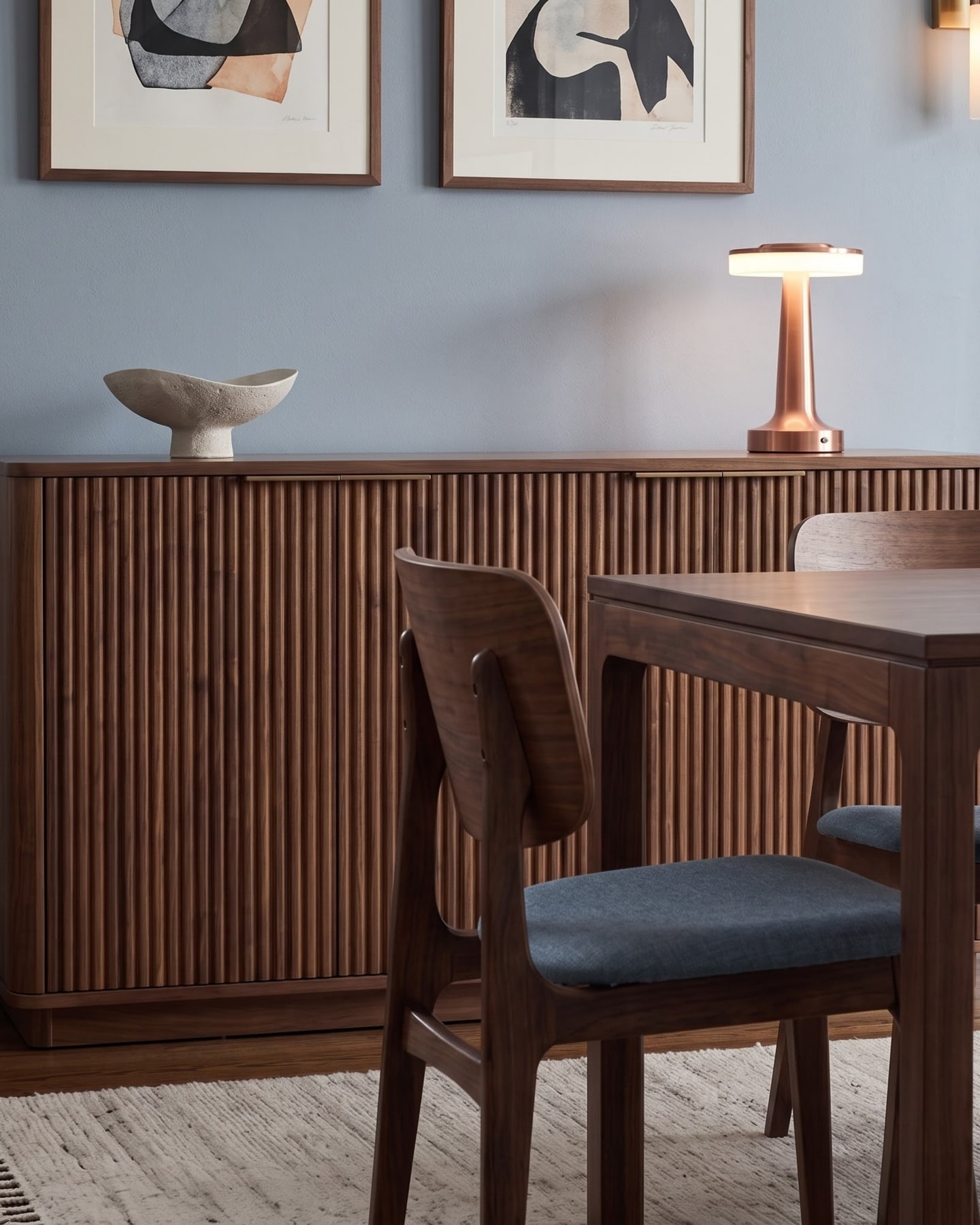 Fluted walnut credenza topped with a modern copper lamp and stone bowl beside a timber dining chair.
