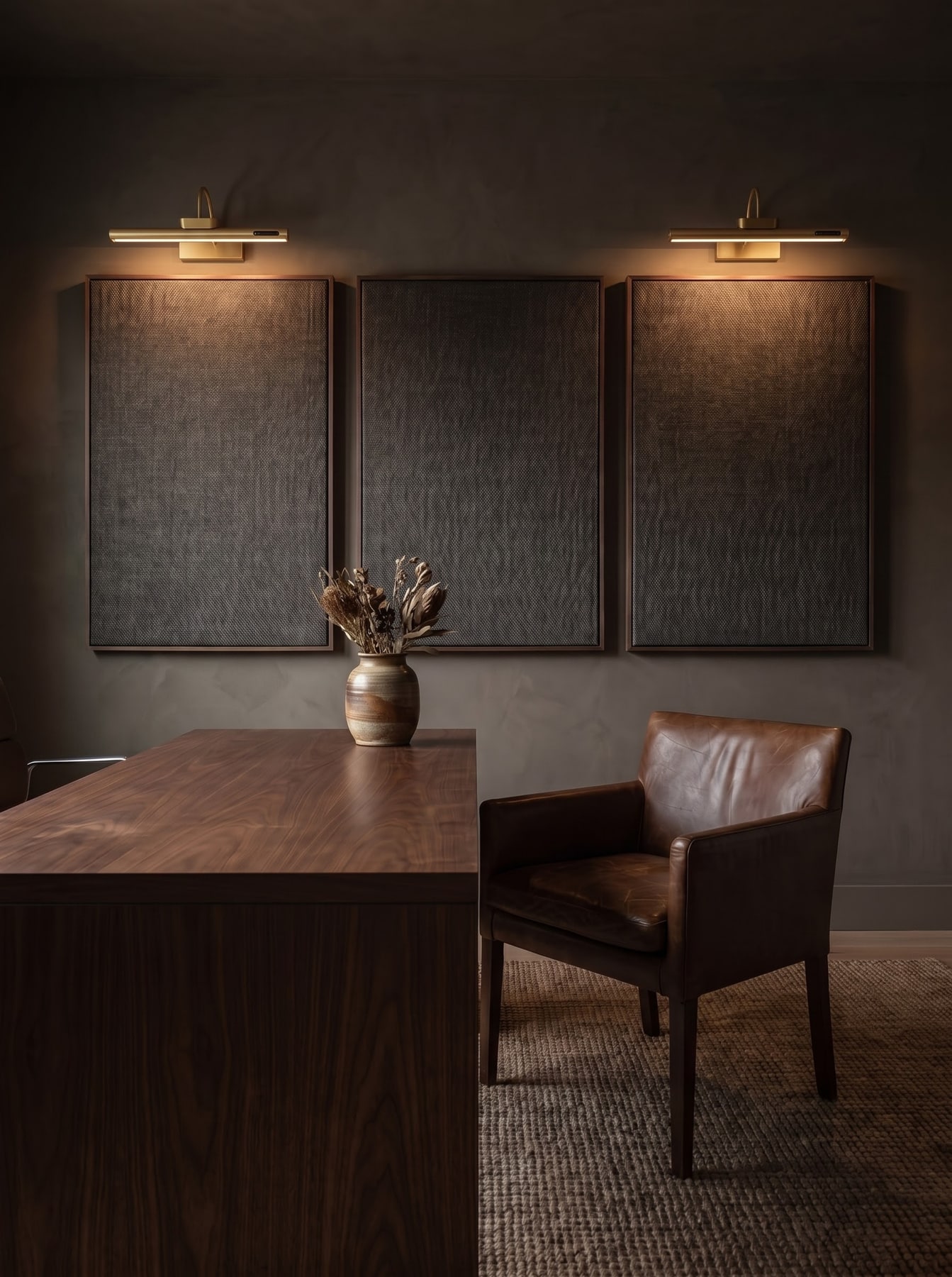 Dark home office with a walnut desk, brown leather armchair, woven rug, and three framed wall panels illuminated by brass picture lights.