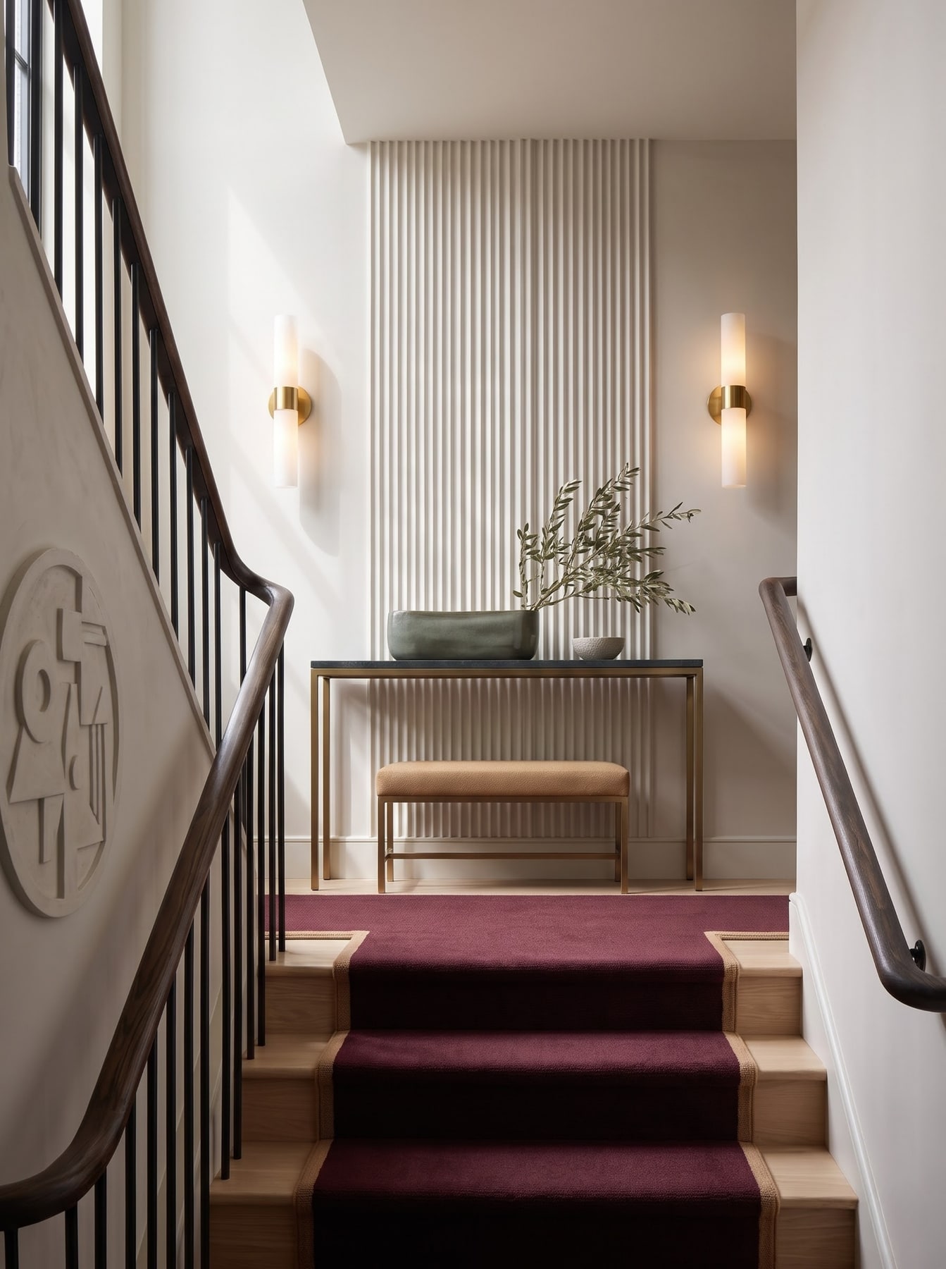 Stair landing with deep red carpet runner, fluted plaster wall panel, slender console table, and two modern tube sconces.