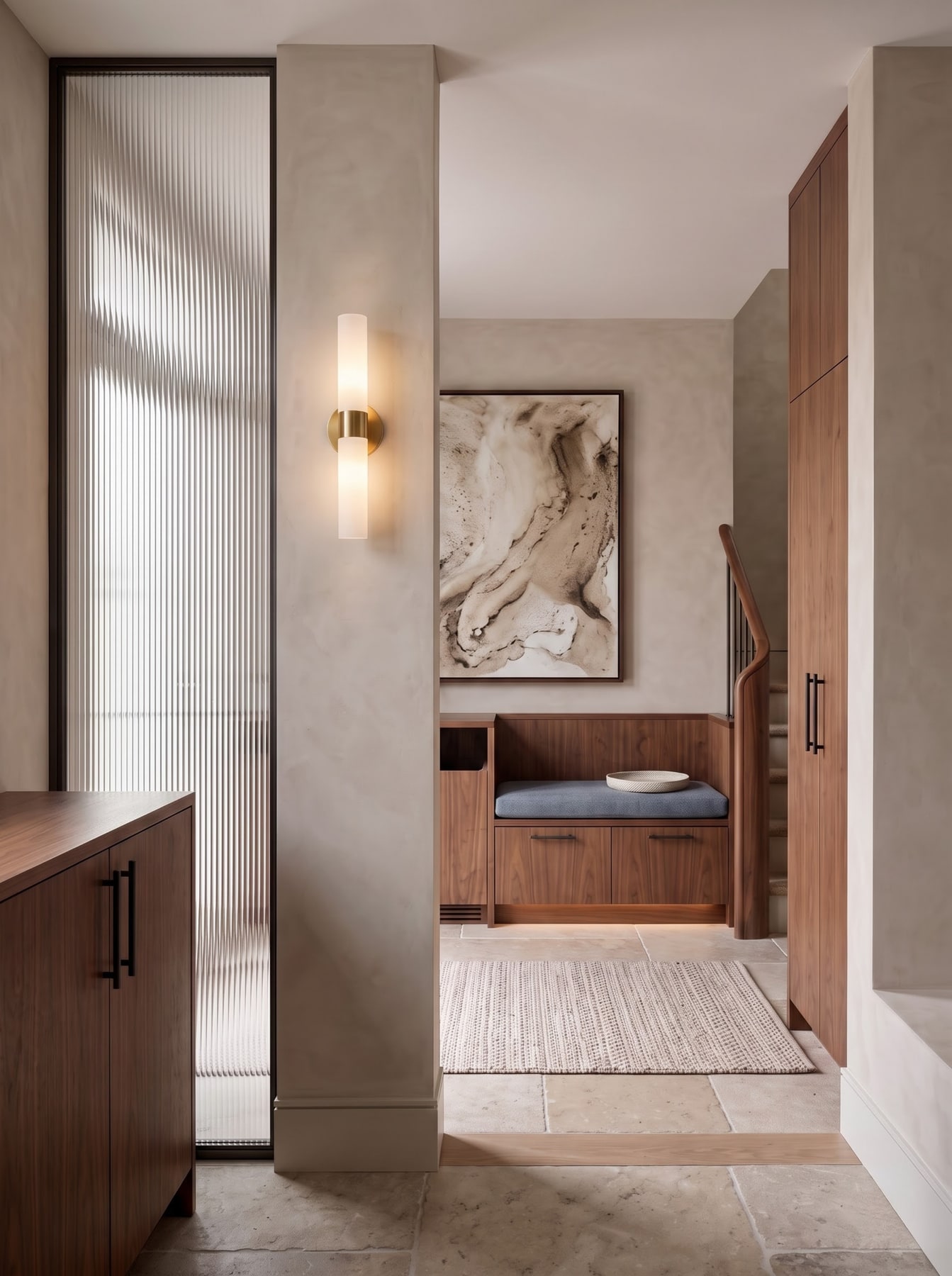 Entry vestibule featuring a ribbed glass screen, walnut bench with blue cushion, brass wall sconce, and limestone tile floor.