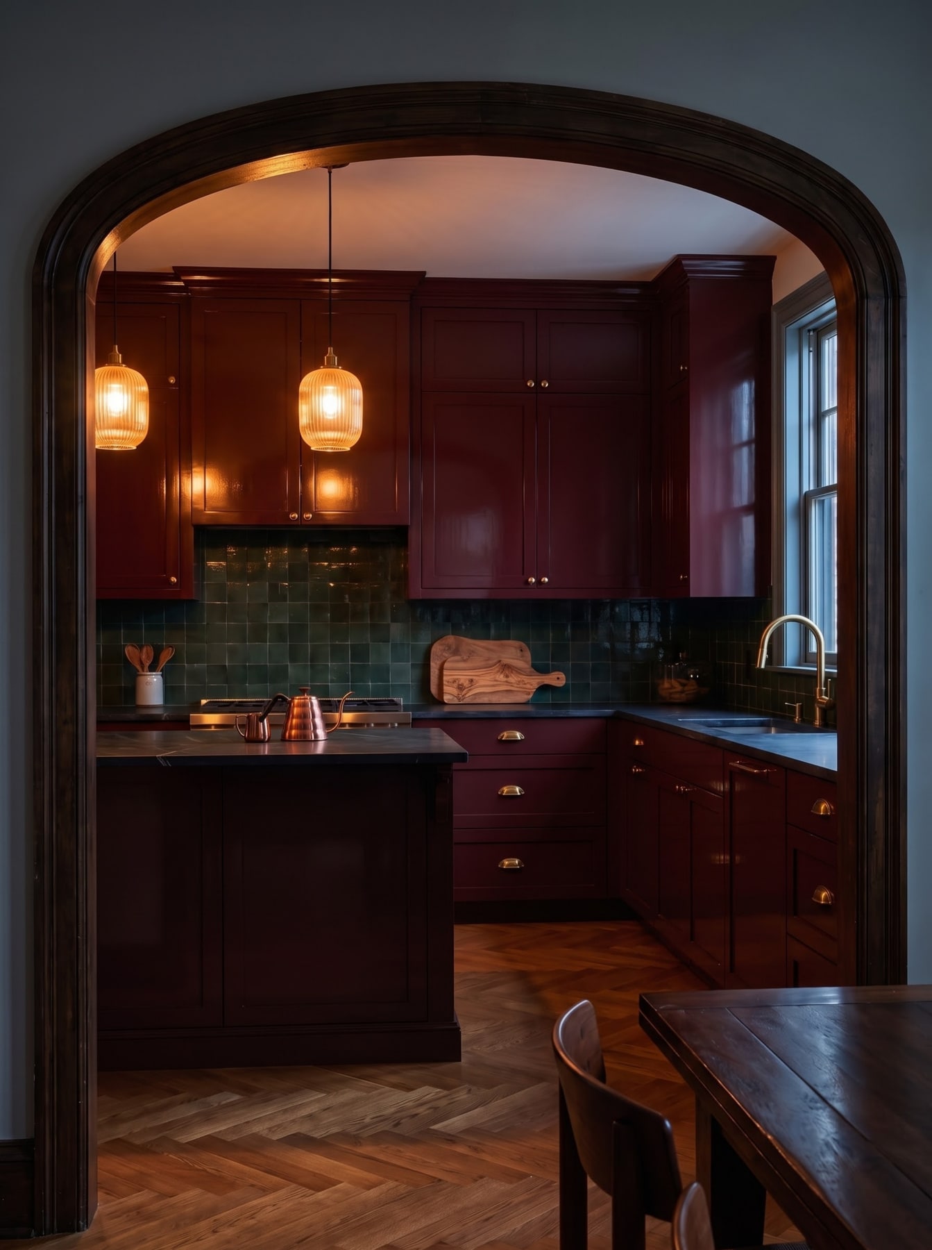 Timber archway framing a deep plum kitchen with glossy green tile backsplash, dark counters, and ribbed amber pendant lights.