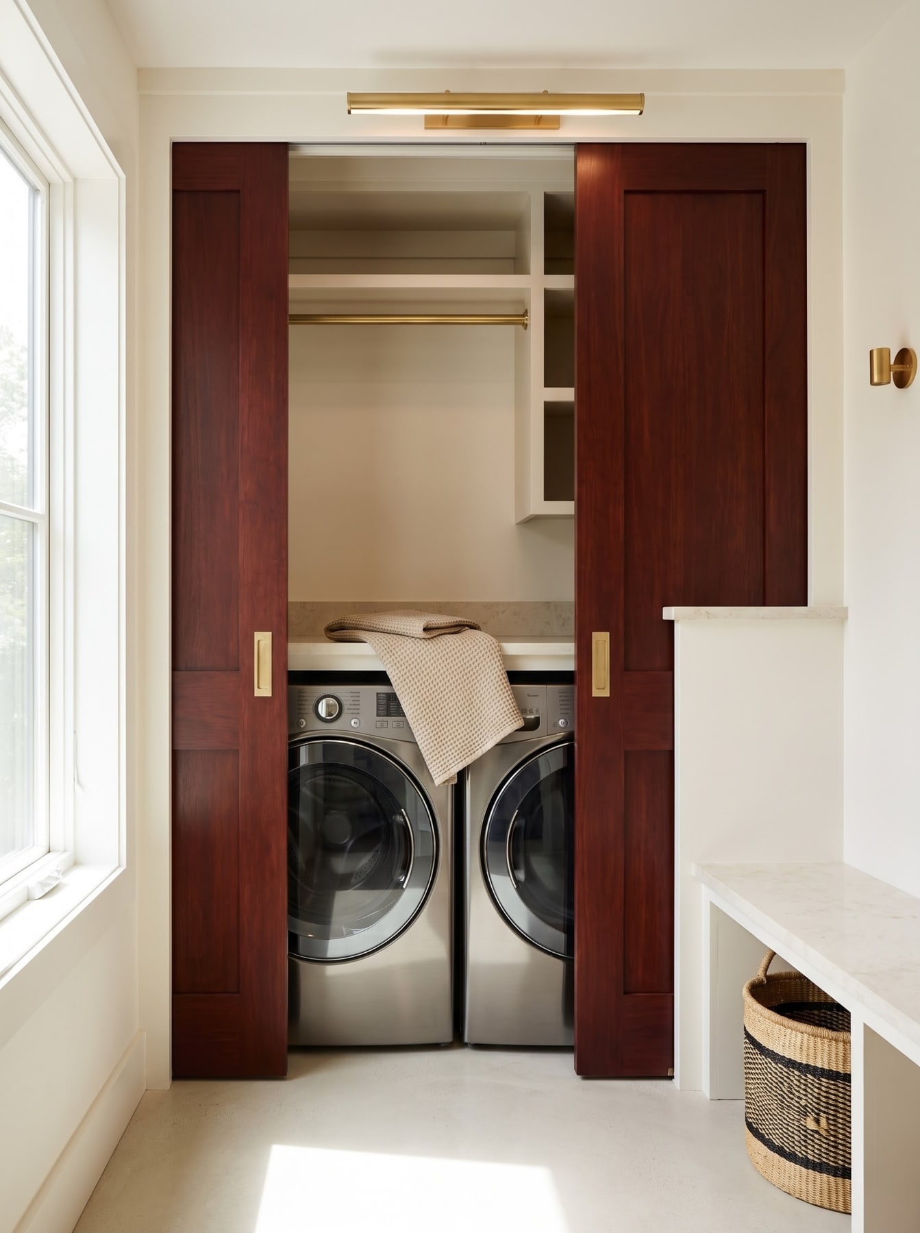 Laundry closet with rich timber pocket doors framing a stacked-counter washer-dryer set, hanging rail, and brass picture light.