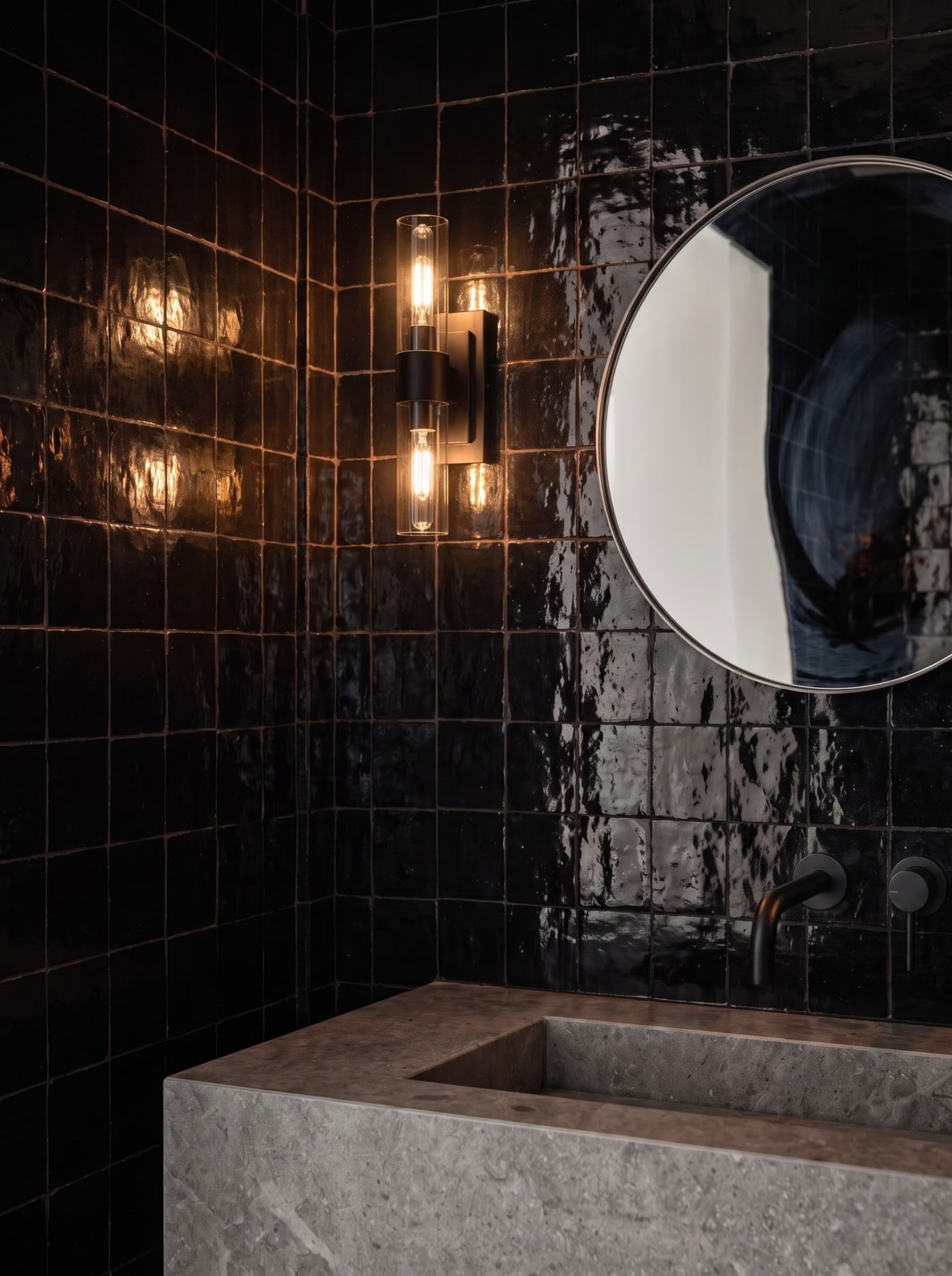 Moody powder room with glossy black zellige tile walls, sculpted grey stone basin, black tapware, and a glowing sconce.