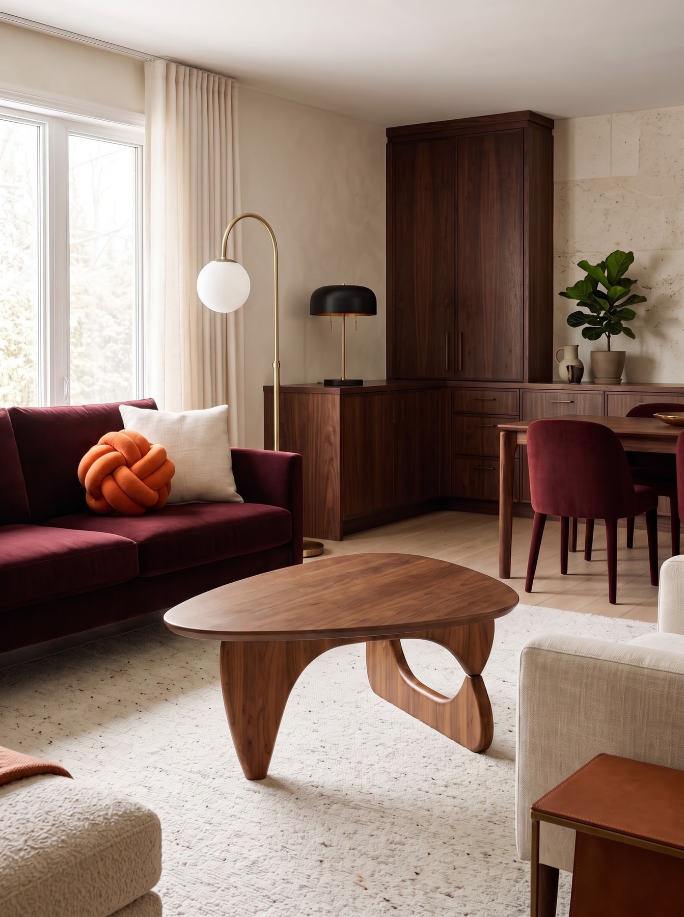 Living space with deep oxblood velvet sofa, sculptural walnut coffee table, curved brass floor lamp, and adjacent dining area.