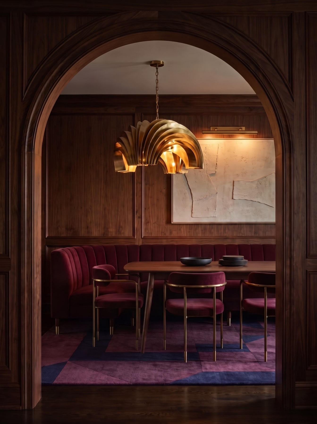 Dark wood archway leading into a dining room with curved burgundy velvet banquette, sculptural brass chandelier, and geometric rug.