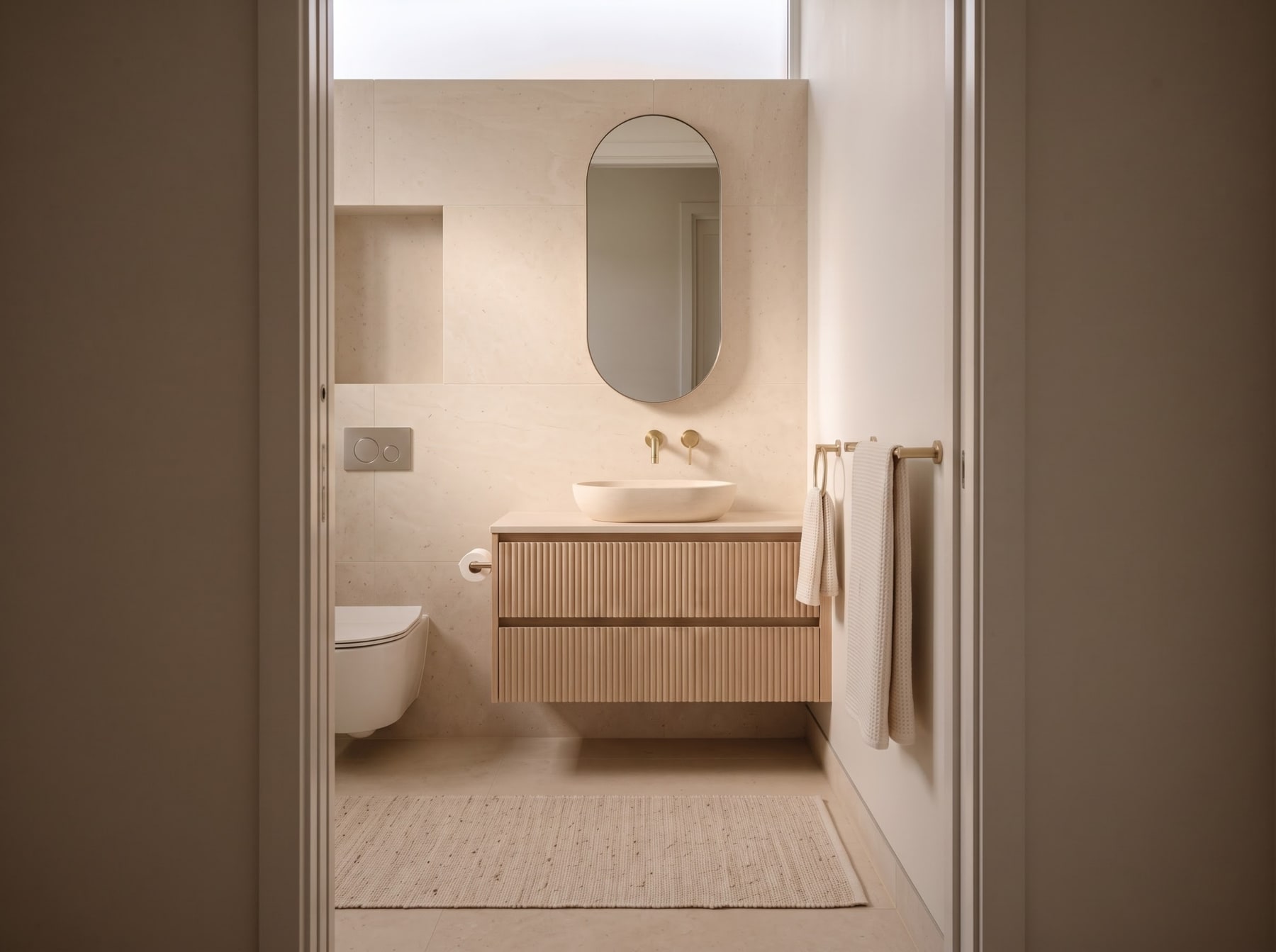Limestone-tiled bathroom with floating fluted oak vanity, vessel sink, wall-mounted brass tapware, pill mirror, and woven floor mat.