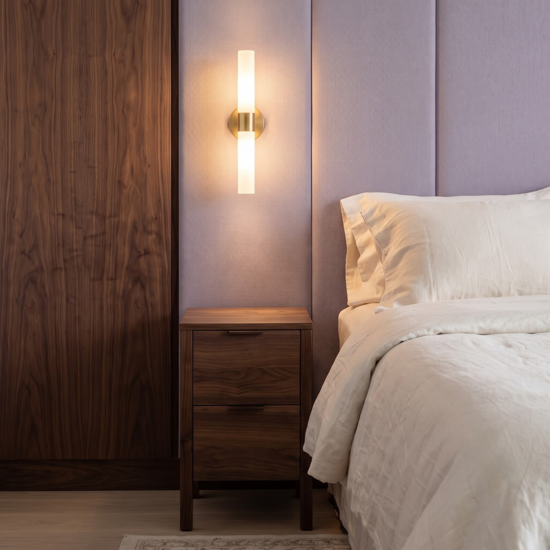 Bedside vignette with a walnut nightstand, illuminated brass cylinder sconce, lilac upholstered wall, and crisp white bed linens.