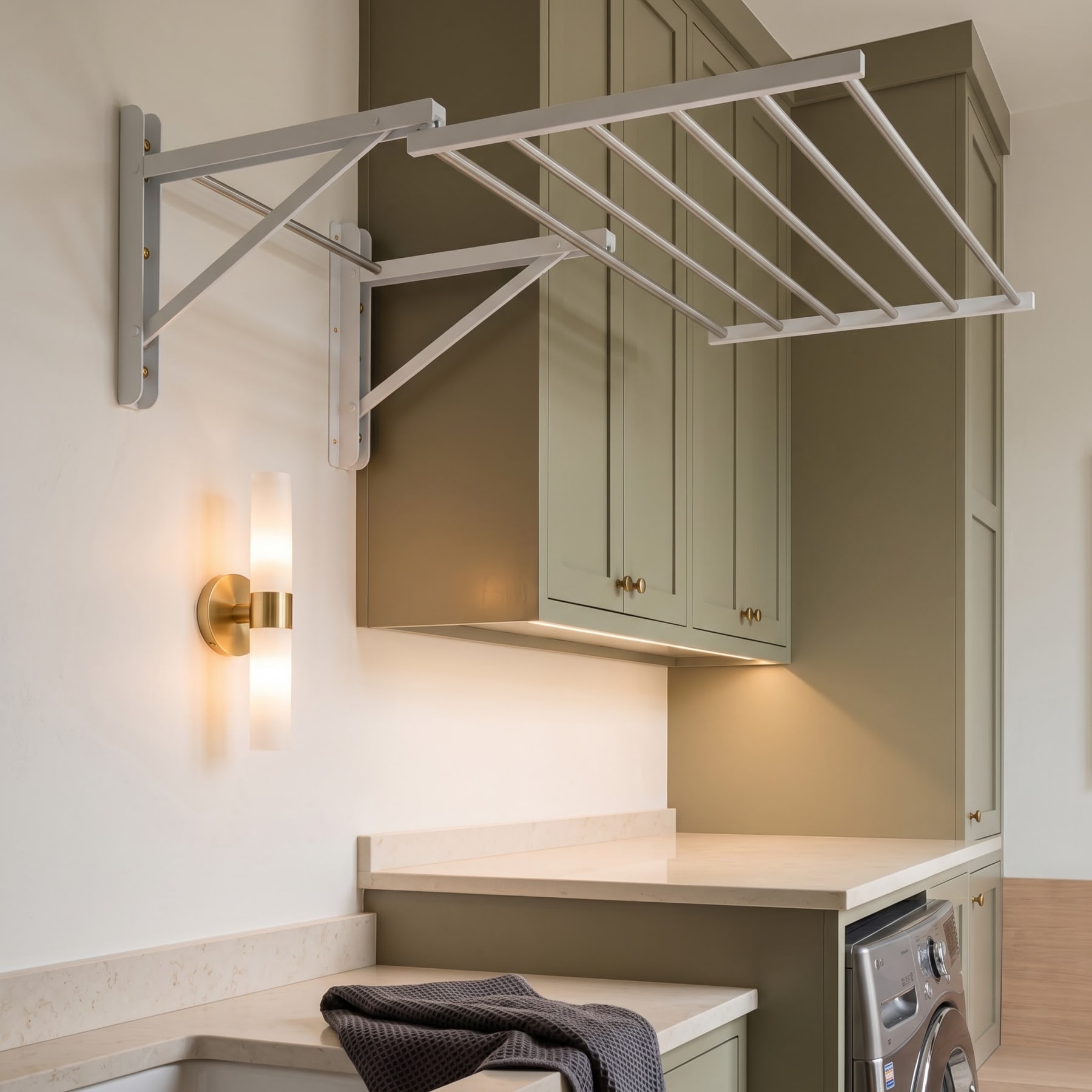 Wall-mounted fold-out drying rack extended above a stone countertop and olive-green cabinetry with brass sconce.