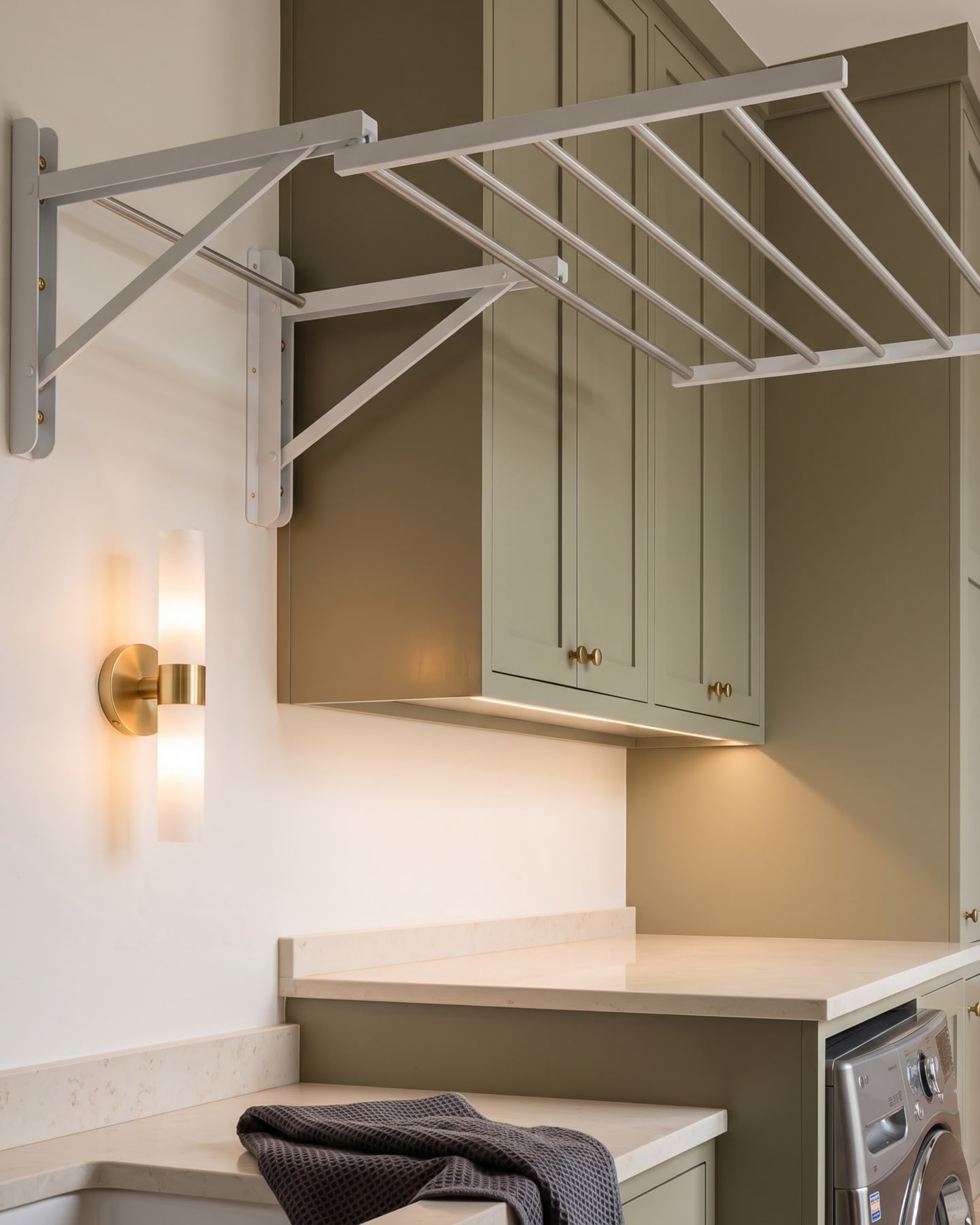 Fold-out wall drying rack extended above a stone countertop and olive-green cabinetry with illuminated brass sconce.